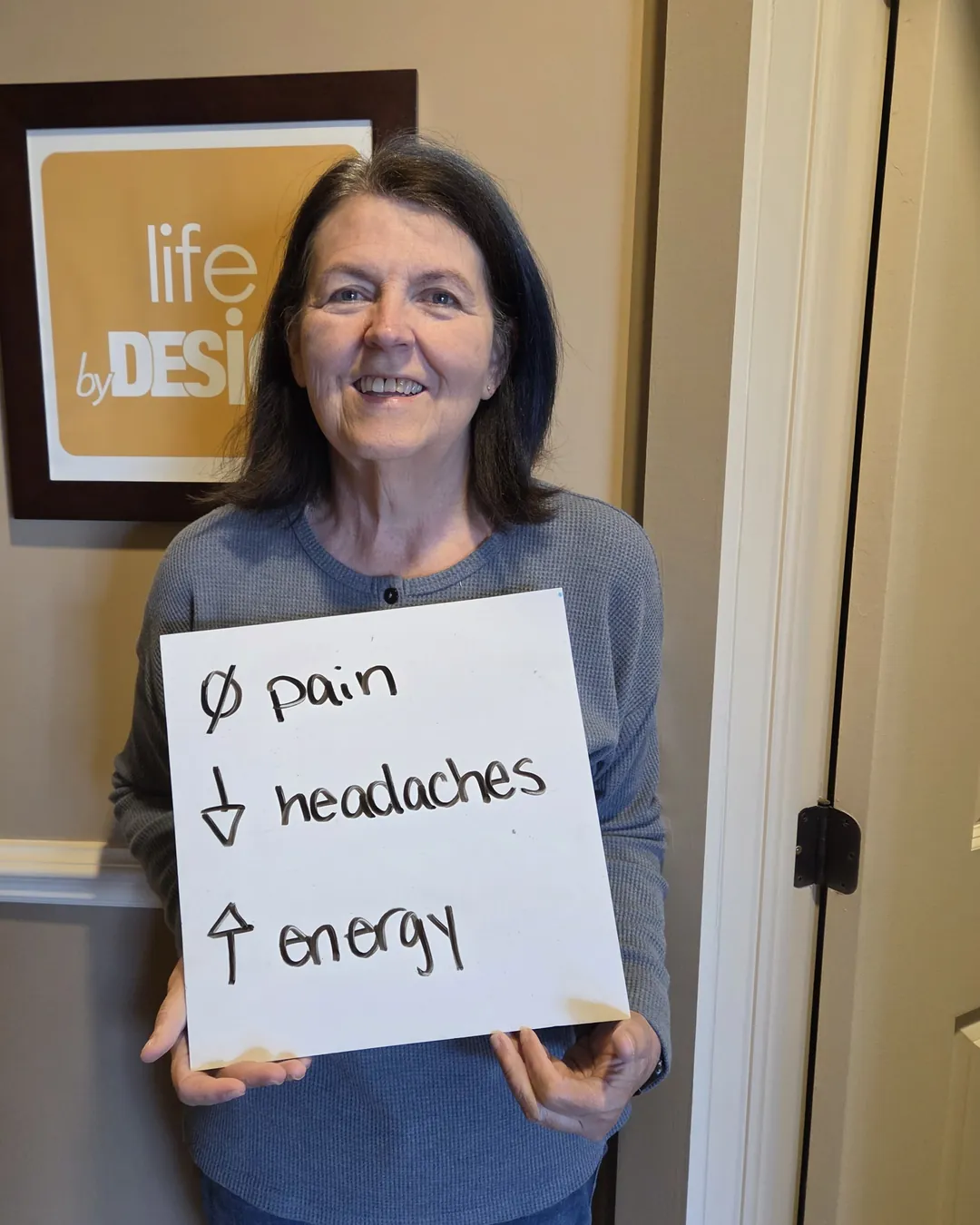 Smiling patient at Synergy Wellness Group in Kanata holding a sign that reads "no pain, fewer headaches, more energy"
